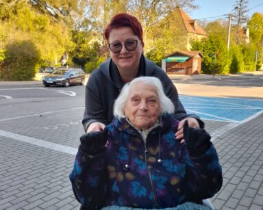 Zbliżenie. Kobieta w szarej bluzie i okularach pozuje do zdjęcia z seniorką na wózku. Seniorka na nogach ma kocyk.