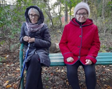 Zbliżenie. Dwie seniorki siedzą na ławeczce. Jedna ma na sobie płaszcz z kapturem, druga czerwoną kurtkę. W tle drzewa.