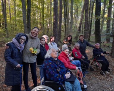 Las. Grupa osób pozuje do zdjęcia grupowego. Wszyscy ubrani w ciepłe kurtki. W tle wysokie drzewa, liście na ziemi.