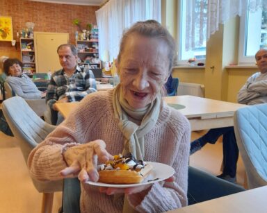 Sala. Seniorka siedzi na fotelu, trzyma w ręku talerzyk. Na talerzyku udekorowany gofr. Kobieta uśmiecha się.