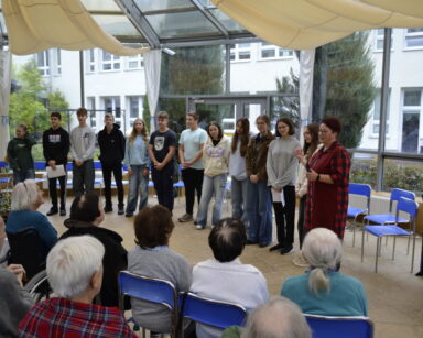 Sala. Grupa młodzieży stoi twarzą zwrócona do seniorów. W tle okna i widok na ogród.