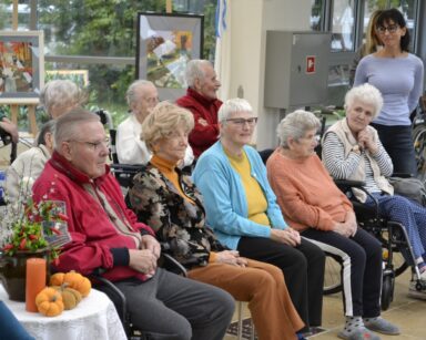 Zbliżenie. Grupa seniorów siedzi na krzesłach. Obok stoi młoda kobieta. W tle na sztalugach stoją obrazy.