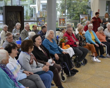 Zbliżenie. Na sali grupa senior siedzi na krzesłach i śpiewa piosenki. W tle okna, Zasłony z sufitu, obrazy na sztalugach.