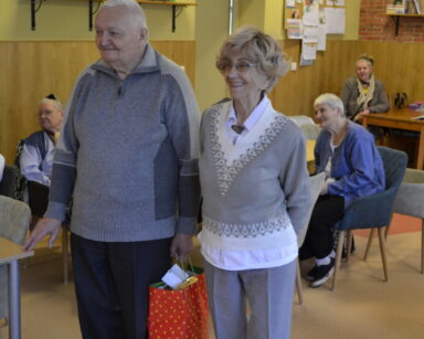 Sala. Para seniorów stoi i uśmiecha się. Senior trzyma czerwoną torbę z prezentami. W tle siedzą przy stole seniorki.