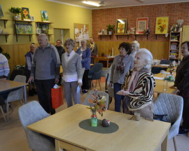 Sala. Seniorzy stoją przy stołach i śpiewają. Senior trzyma czerwoną torbę z prezentami, seniorka kartkę.