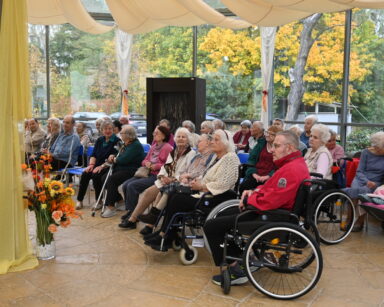 Sala. Spora grupa seniorów siedzi na krzesłach lub wózkach. W tle kotary, szklane duże okna, widok na parking.