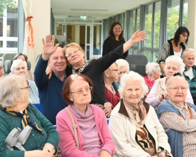 Zbliżenie. Grupa radosnych seniorów pozuje do zdjęcia. Do jednego seniora przytula się uśmiechnięta kobieta i macha ręką.