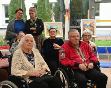 Zbliżenie. Sala. Grupa seniorów siedzi na krzesłach, za nimi stoją trzy osoby. W tle wystawa obrazów, za oknem ogród.