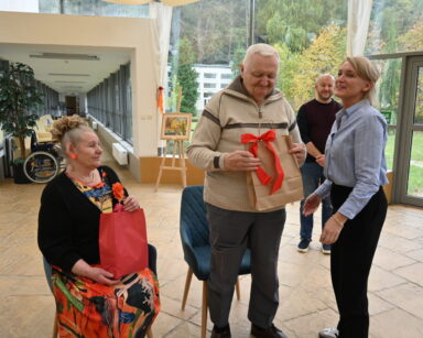 Zbliżenie. Kobieta stoi przy seniorze który trzyma w rękach torbę z niespodzianką, obok siedzi seniorka, też trzyma torbę.