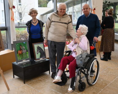 Zbliżenie. Uśmiechnięta seniorka na wózku ściska dłoń seniorowi. Za kobietą stoi senior, przy obrazach uśmiechnięta seniorka.