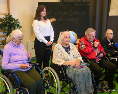 Scena. Grupa seniorów siedzi na krzesłach i oglądają przypięte medale dla nauczycieli. W tle stół, globus, mapa i tablica.