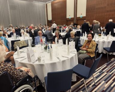 Sala. Okrągłe stoły. Goście. Każdy ubrany w wieczorowy strój. Na stołach podarki od Ziaja, zastawa, kwiaty w wazonach.