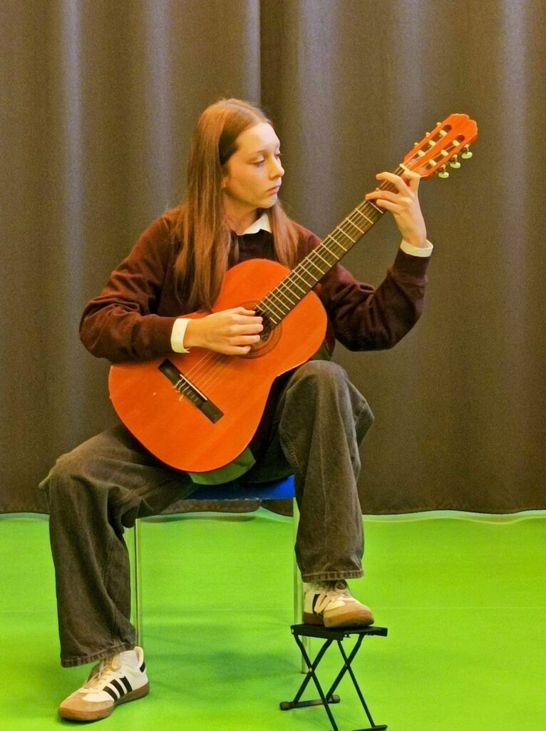 Zbliżenie. Młoda dziewczyna, ubrana w sweter i długie spodnie, siedzi na krześle i trzyma w ręku gitarę.