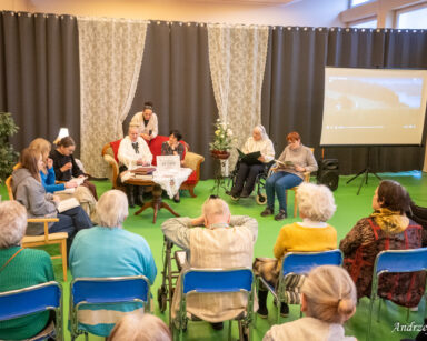 Sala. Grupa osób. Na fotelach siedzą seniorki i młodzież. Czytają książki. W tle kotary i firanki