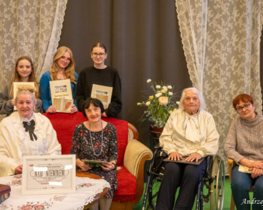 Zbliżenie. Cztery seniorki i trzy młode dziewczyny pozują do zdjęcia. W tle szare kotary i koronkowe firany.