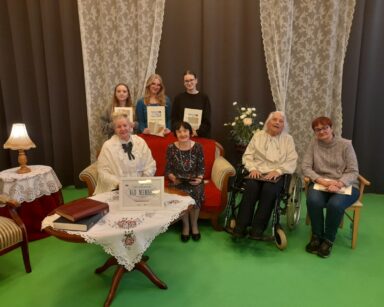 Zbliżenie. Cztery seniorki i trzy młode dziewczyny pozują do zdjęcia. W tle szare kotary i koronkowe firany.