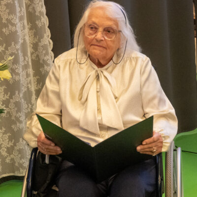 Zbliżenie. Seniorka w białej koszuli i ciemnych spodniach siedzi na wózku. W rękach trzyma zielona okładkę.