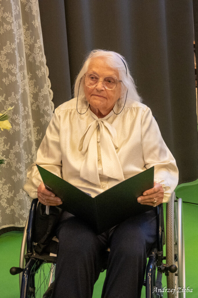 Zbliżenie. Seniorka w białej koszuli i ciemnych spodniach siedzi na wózku. W rękach trzyma zielona okładkę.