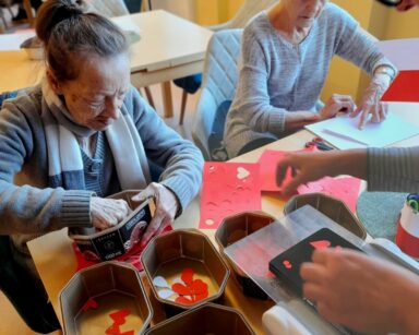 Sala. Kobieta w koszuli w paski podaje seniorkom kolorowe wycinanki. Seniorki wyciągają białe i czerwone serca.