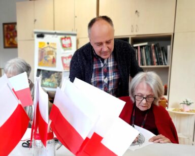 Sala. Mężczyzna pochyla się nad seniorką w czerwonym swetrze. Kobieta koloruje na kartce orła. Obok stoją flagi Polski.