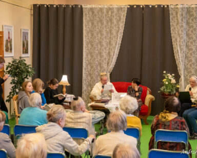 Sala. Grupa osób. Na fotelach siedzą seniorki i młodzież. Czytają książki. W tle kotary i firanki.