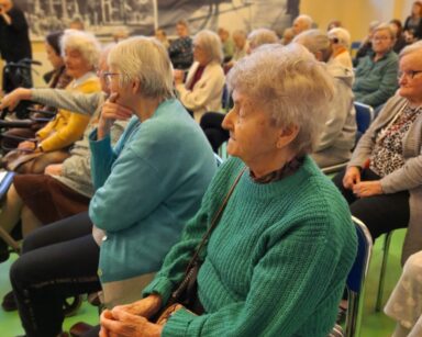 Sala. Na sali pełno seniorów siedzi na krzesłach. Zwróceni są w jedną stronę. W tle czarnobiałe zdjęcia na ścianach.