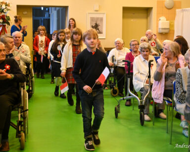 Sala. na sali siedzą seniorzy i klaszczą w dłonie. Do sali wchodzi grupa maluchów z flagami Polski w rączkach.