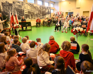 Sala. Na sali siedzą seniorzy. Na scenie siedzą maluchy z flagami Polski w dłoniach. Przy mikrofonie stoi seniorka.