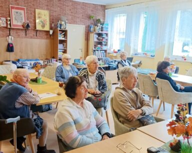 Sala. Grupa seniorek siedzi przy stołach i patrzy w jedną stronę. Na stołach jesienne kompozycje w wazonikach.