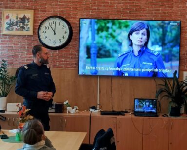 Sala. Na monitorze wyświetlona pani policjant. Obok monitora stoi pan policjant. W tle przy stołach siedzą seniorki.