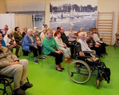 Sala. Seniorzy siedzą na krzesłach i oglądają występ muzyków. W tle na ścianie czarno białe zdjęcia.