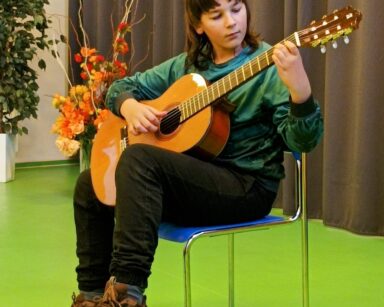 Zbliżenie. Młody chłopak w długich włosach, ciemnej bluzie i długich spodniach siedzi na krześle. Trzyma w rękach gitarę.