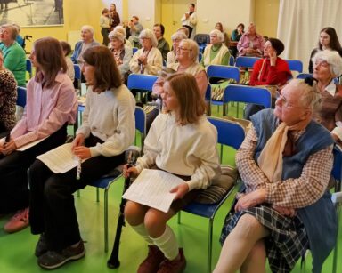 Sala. Na sali siedzą seniorzy i młodzi ludzie , którzy trzymają w rękach instrumenty muzyczne.