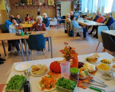 Sala. Przy stołach siedzą seniorzy. Na przeciwko seniorów stoi stolik z produktami do pasty na kanapki.