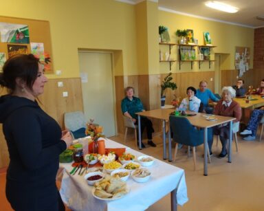 Sala. Kobieta w czarnej sukience stoi przy stole , na którym są produkty do pasty na kanapki. Na sali siedzą seniorzy.