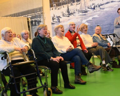 Sala. Grupa radosnych seniorów klaszcze i śpiewa. Obok przy ścianie stoi kobieta i uśmiecha się do seniorów.