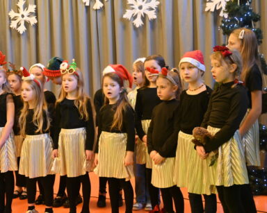 Zbliżenie. Grupa dziewczynek w złotych spódniczkach, czarnych bluzeczkach i czapkach Mikołaja śpiewa na scenie.