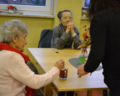 Zbliżenie. Dwie seniorki siedzą przy stoliku. Jedna z seniorek miesza herbatę, koło niej pochyla się kobieta w czerni.