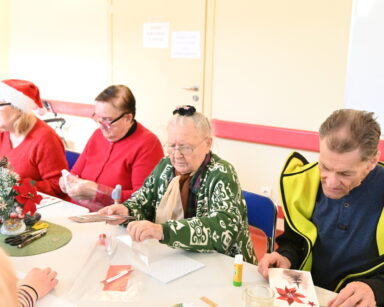 Zbliżenie. Grupa seniorów siedzi przy stole. Seniorka trzyma w ręku kartkę, obok senior ogląda kartkę z choinką.