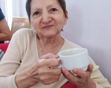 Na czerwonym fotelu siedzi seniorka w beżowym sweterku. W dłoniach trzyma białą filiżankę z kawą i uśmiecha się patrząc w obiektyw