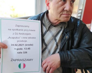 Na tle okna siedzi senior w skórzanej czarnej kurtce i szarym swetrze w romby. W rękach trzyma białą ulotkę z zaproszeniem do udziału w spotkaniu przy włoskiej muzyce. Na ulotce zamieszczone jest kółko z pionowymi pasami kolorów: zielonym, białym, czerwonym. Symbolizują one włoską flagę