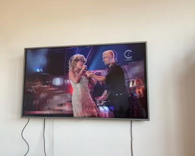 Na jasnej ścianie wisi płaski telewizor. W zatrzymanym ujęciu widać jak włoski piosenkarz Eros Ramazzotti śpiewa w duecie z amerykańską piosenkarką Tiną Turner