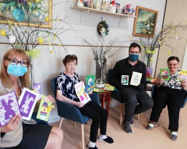 Zaczynając od lewej, na krzesłach siedzą: dyrektor Agnieszka Cysewska, seniorka, administrator Łukasz Cielebański, seniorka. Wszyscy się uśmiechają. W rękach trzymają wielkanocne kartki z życzeniami. W tle widać świąteczne dekoracje, wazony z gałązkami leszczyny ozdobione pisankami, piórkami i wstążkami