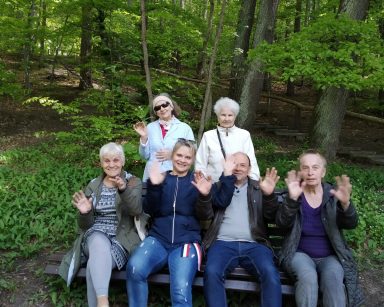 Zielony las. Terapeutka Beata Gadomska i seniorzy na ławce. Za nimi dwie seniorki. Wszyscy machają.