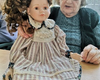 Seniorka trzyma lalkę. Lalka ma brązowe, kręcone włosy. Jest ubrana w sukienkę z kołnierzem i pasisty fartuszek.
