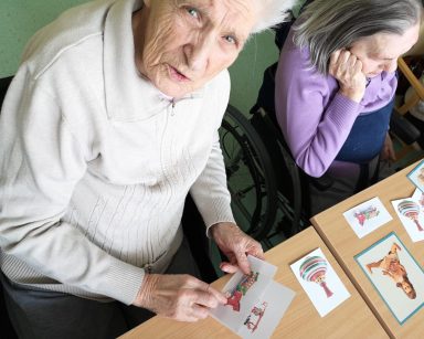 Dwie seniorki siedzą przy stole i oglądają rozłożone obrazki. Na obrazkach drewniane zabawki, kolorowe bączki, lalki.