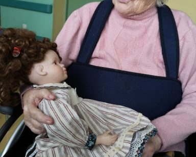 Seniorka trzyma na kolanach lalkę. Lalka ma brązowe, kręcone włosy. Jest ubrana w sukienkę z kołnierzem i pasisty fartuszek.
