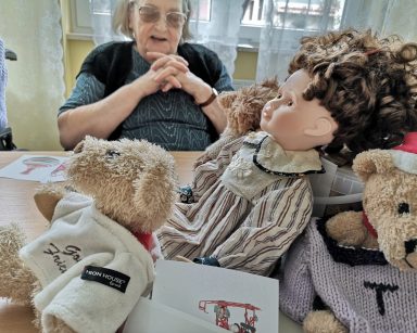 Przy stole siedzi uśmiechnięta seniorka. Przed nią na blacie zabawki: lalka, misie.