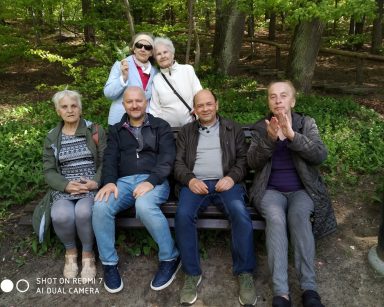 Zielony las. Kierownik Arkadiusz Wanat i seniorzy na ławce. Za nimi dwie seniorki.