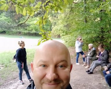 Kierownik Arkadiusz Wanat uśmiecha się do zdjęcia. Na skraju lasu seniorzy na ławkach, obok terapeutka Beata Gadomska.